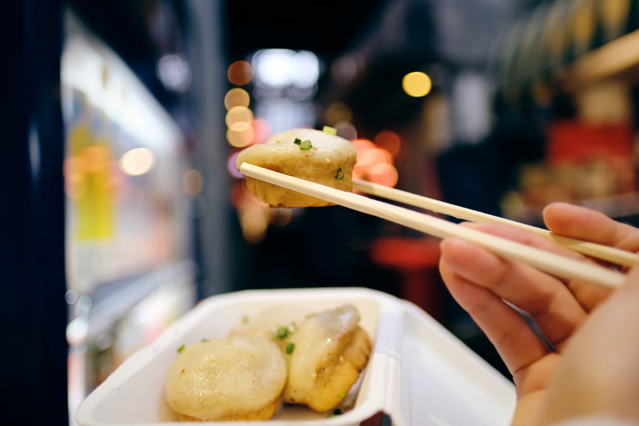 A hand uses chopsticks to pick up a steamed bun from a white container, with blurred city lights and street market background.