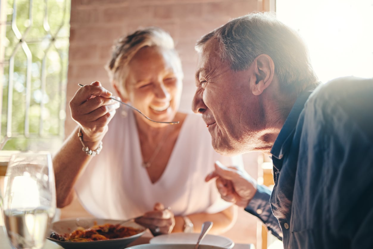 An older woman smiling and feeding pasta to an older man with a spoon at a sunlit table, both appearing happy and enjoying their meal together.