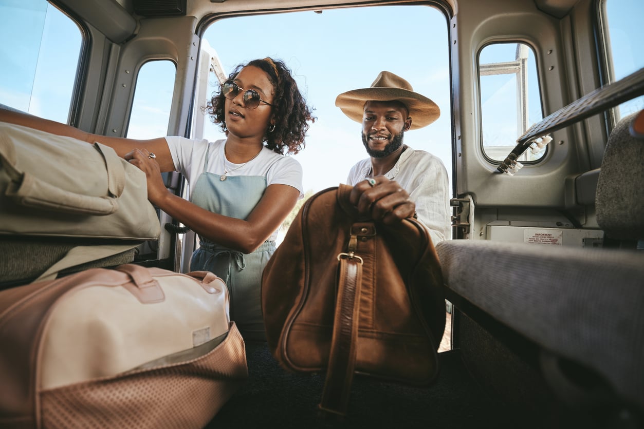A woman and a man load travel bags into the back of a van on a sunny day, preparing for a trip reminiscent of travel in the 60s and 70s.
