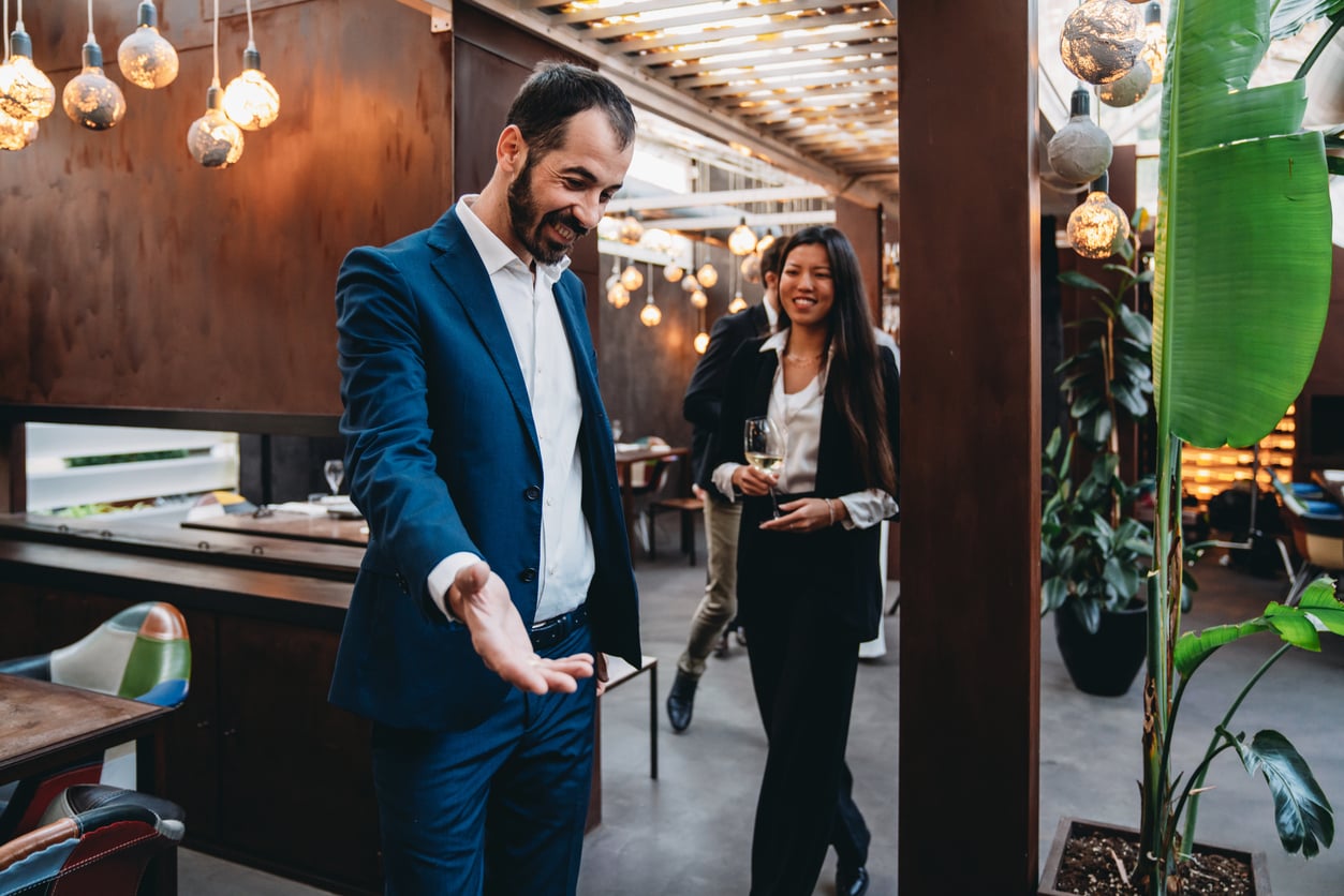 A man in a blue suit gestures with his hand while smiling, leading a woman in business attire who is holding a glass of wine in a modern, warmly lit restaurant or cafe.