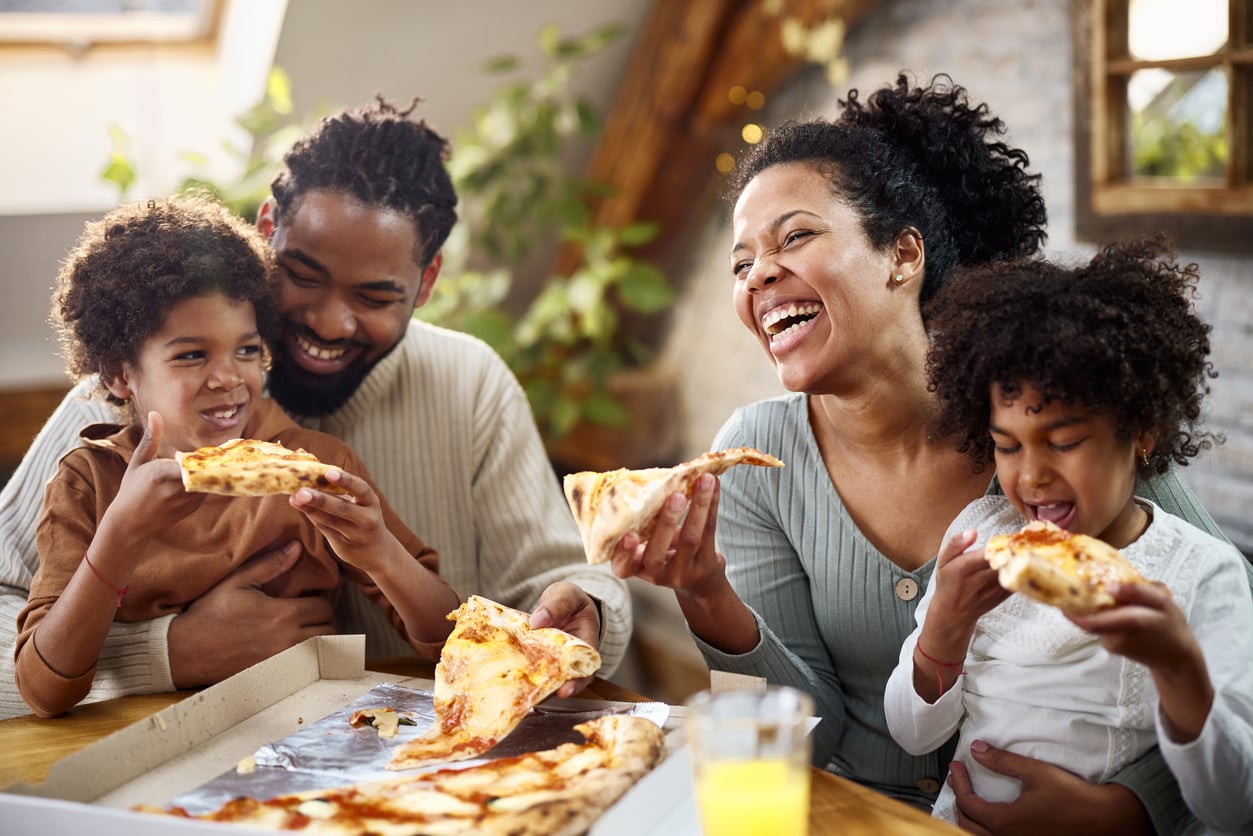 A family of four sits at a table, smiling and laughing while eating pizza together. A pizza box and a glass of juice are in front of them, and they appear to be enjoying a fun, casual meal at home.