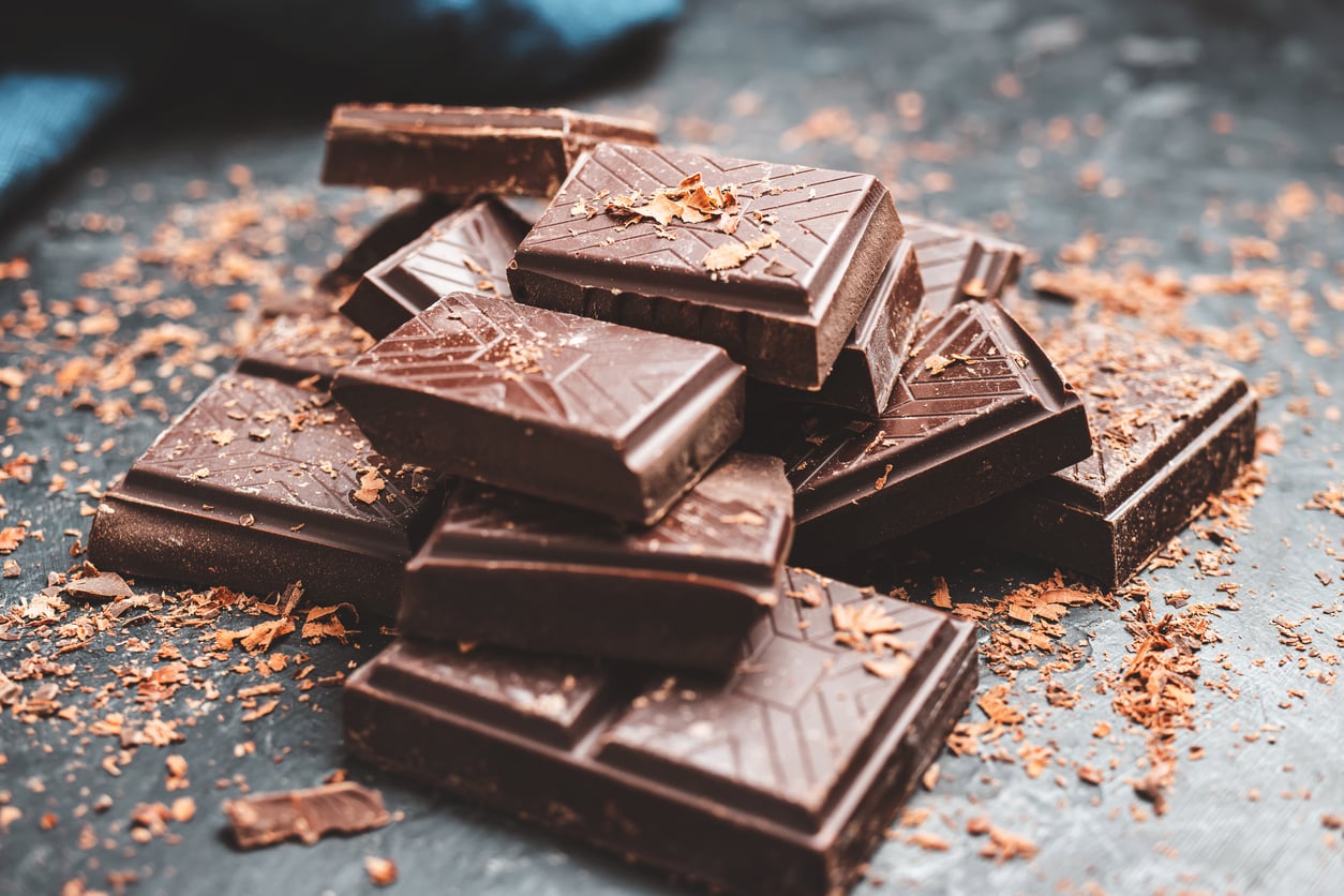 A pile of dark chocolate pieces with geometric patterns, surrounded by chocolate shavings on a dark surface—one of those Foods People Learn to Appreciate Later in Life.