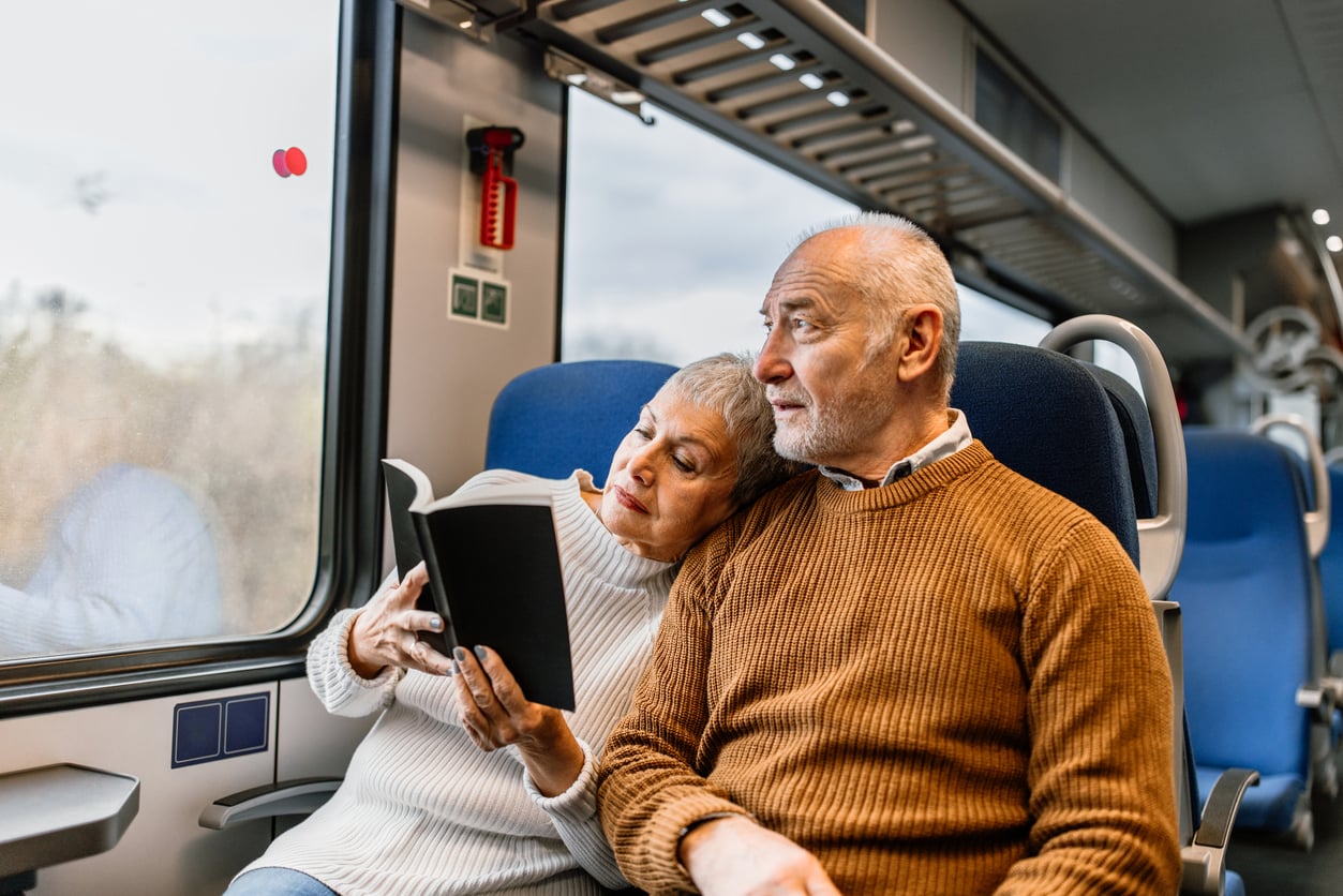 An elderly woman rests her head on a man’s shoulder while reading a book together on a train. Both look out the window, sitting closely on blue seats, with natural light coming in.