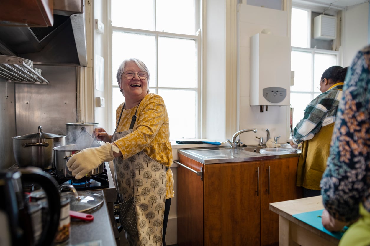 An older woman in an apron smiles while stirring a pot on the stove in a bright kitchen, with two other people preparing food nearby.