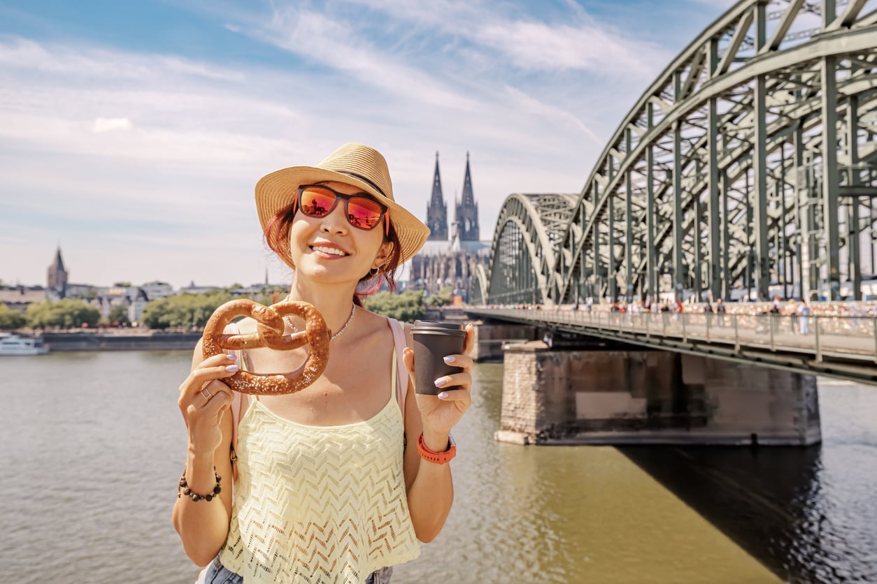 Smiling woman in a hat and sunglasses holds a pretzel and a cup by a river, with a large bridge and Cologne Cathedral in the background under a blue sky.