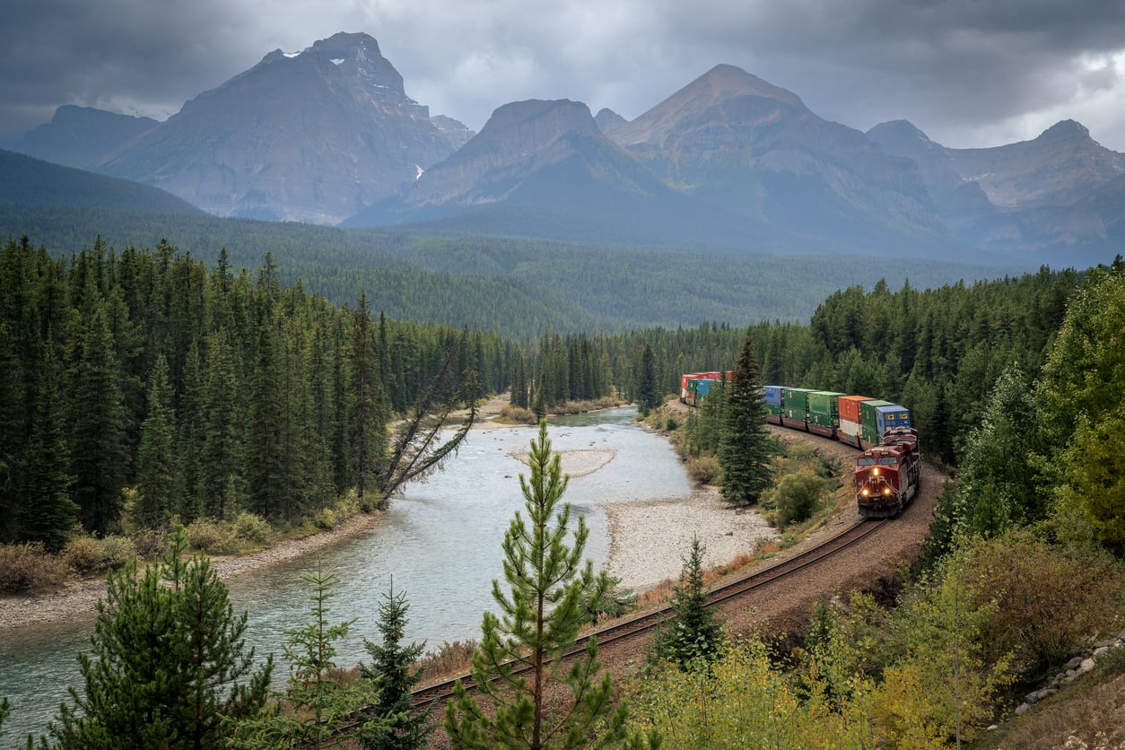 A freight train travels along a curved track beside a river, surrounded by dense pine forests and mountains under a cloudy sky.
