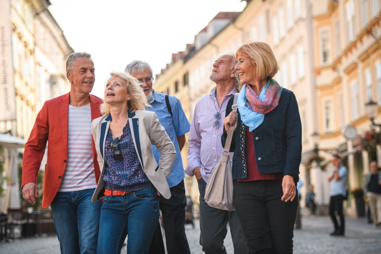 Five older adults walk together down a city street, smiling and talking. The background shows tall, colorful buildings and a sunny atmosphere.