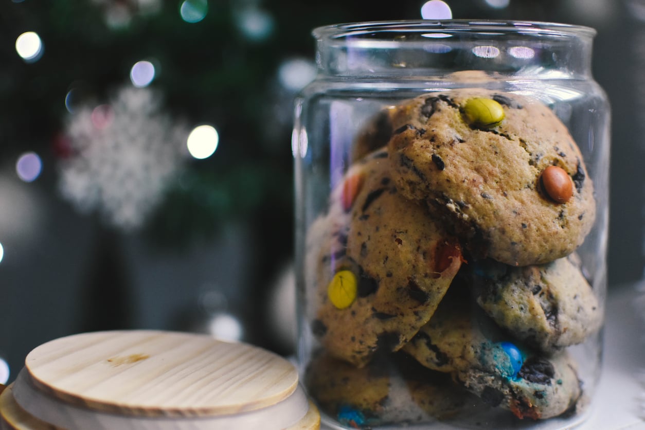 A glass jar filled with chocolate chip cookies topped with colorful candy-coated chocolates, with the jar lid resting nearby and blurred festive lights in the background.