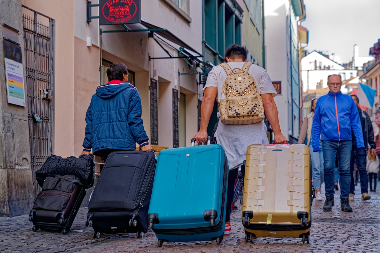 Two people pull large suitcases down a cobblestone street in a city, while other pedestrians walk nearby. The focus is on travel and movement.