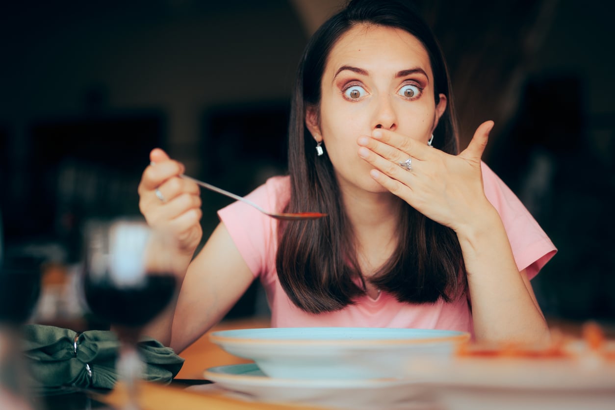 A woman with wide eyes covers her mouth with one hand while holding a spoon in the other, appearing surprised at the dining table.