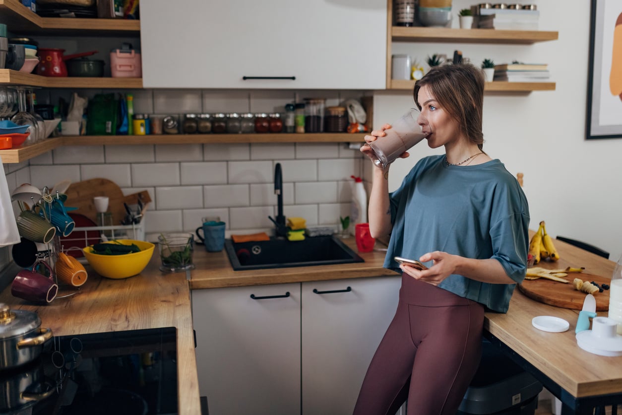 A woman in workout clothes stands in a kitchen, holding a phone and drinking a smoothie. There are fruits, bowls, and cups on the wooden counters and shelves around her.