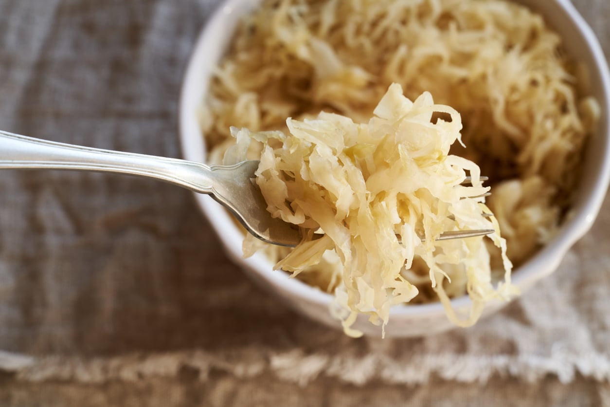 A close-up of a fork holding sauerkraut above a white bowl filled with more sauerkraut on a textured fabric surface—a perfect example of one of those foods people learn to appreciate later in life.