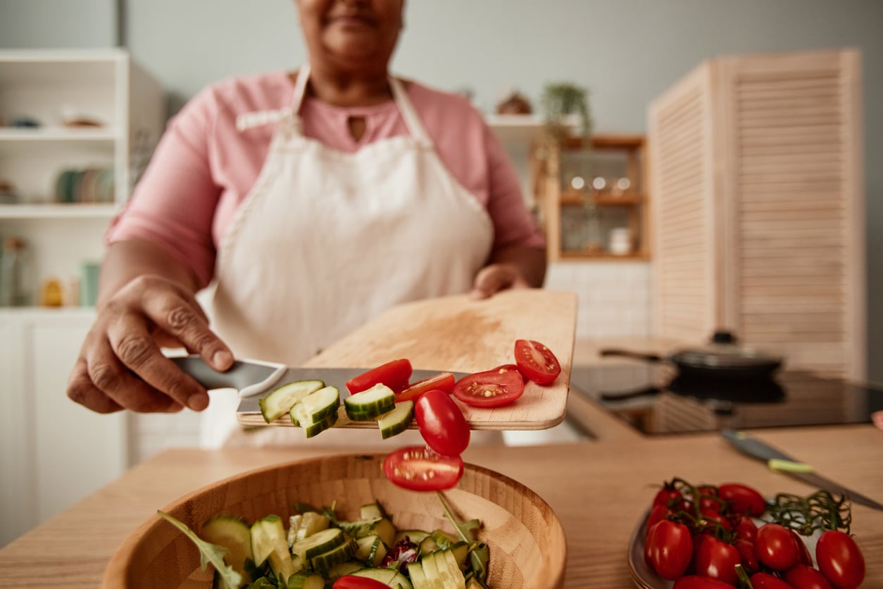 A person in an apron adds sliced cucumbers and cherry tomatoes from a wooden cutting board into a salad bowl in a kitchen.