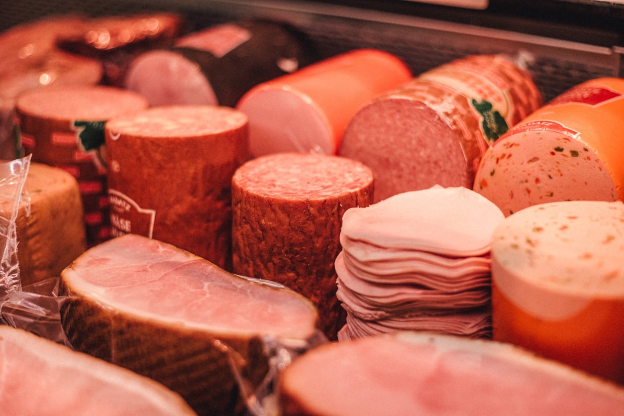 A close-up view of various deli meats, including sliced ham, salami, bologna, and other cold cuts, neatly arranged in a display case at a deli or grocery store.