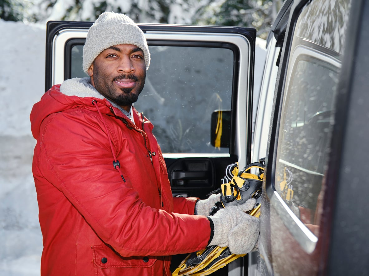 A man in a red jacket, gray hat, and gloves stands beside a vehicle door, holding a pair of yellow snowshoes. Snow and trees are visible in the background.