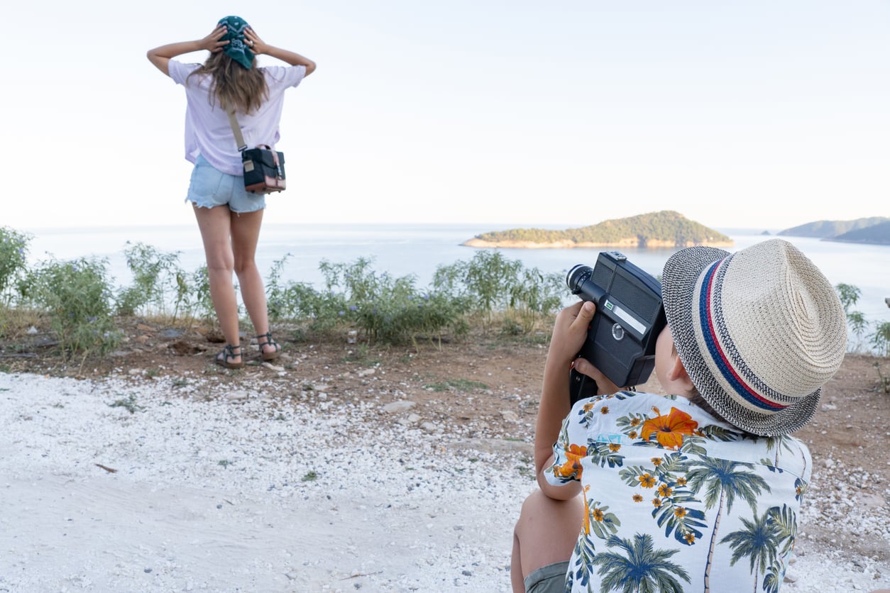 A child wearing a straw hat films a woman standing by the seaside, facing away, capturing a perfect vacation moment. The woman holds her hat with both hands as she enjoys the gentle breeze. The background shows calm water and distant islands under a clear sky.