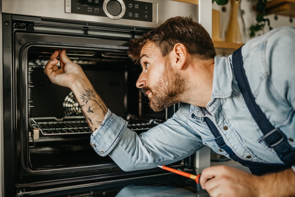 A man in a denim shirt inspects the inside of an oven, holding a screwdriver in one hand and reaching inside with the other. He has a tattoo on his forearm and appears focused on repairing the appliance.
