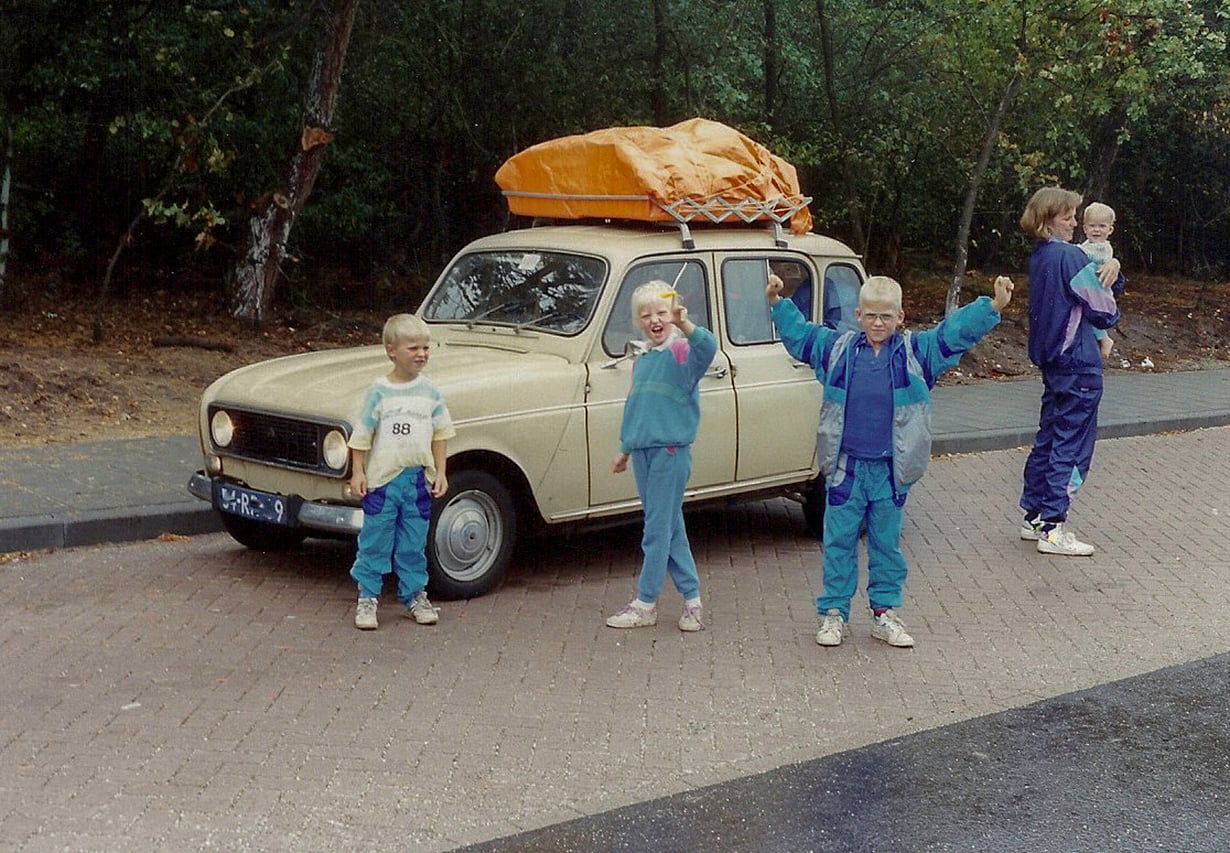 Three kids in colorful tracksuits pose and smile in front of an old beige car with orange luggage on top, capturing the spirit of travel in the 60s and 70s. Behind them, an adult holding a child stands on the sidewalk, with trees and greenery creating a nostalgic backdrop.