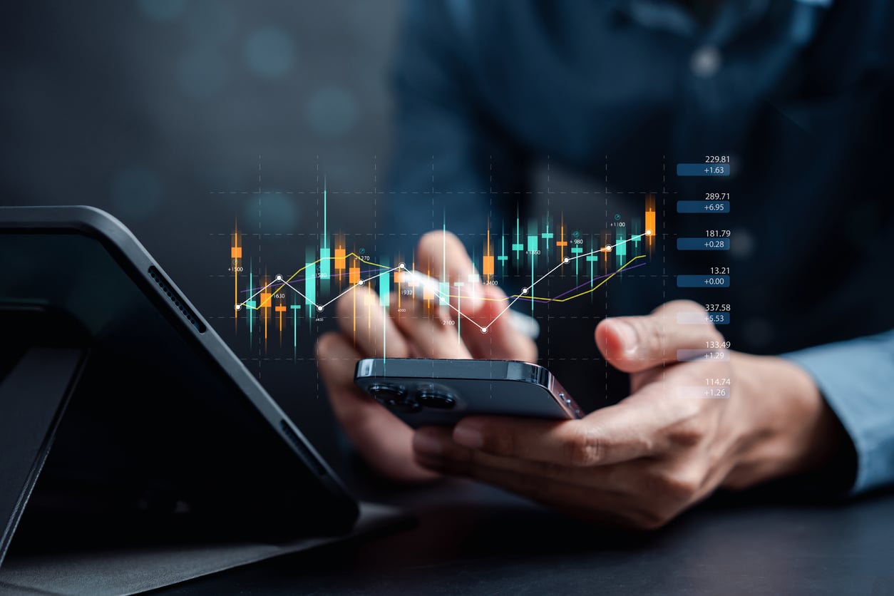 A person uses a smartphone and stylus near a tablet, with digital stock market charts and candlestick graphs overlaid, representing online trading or financial analysis.