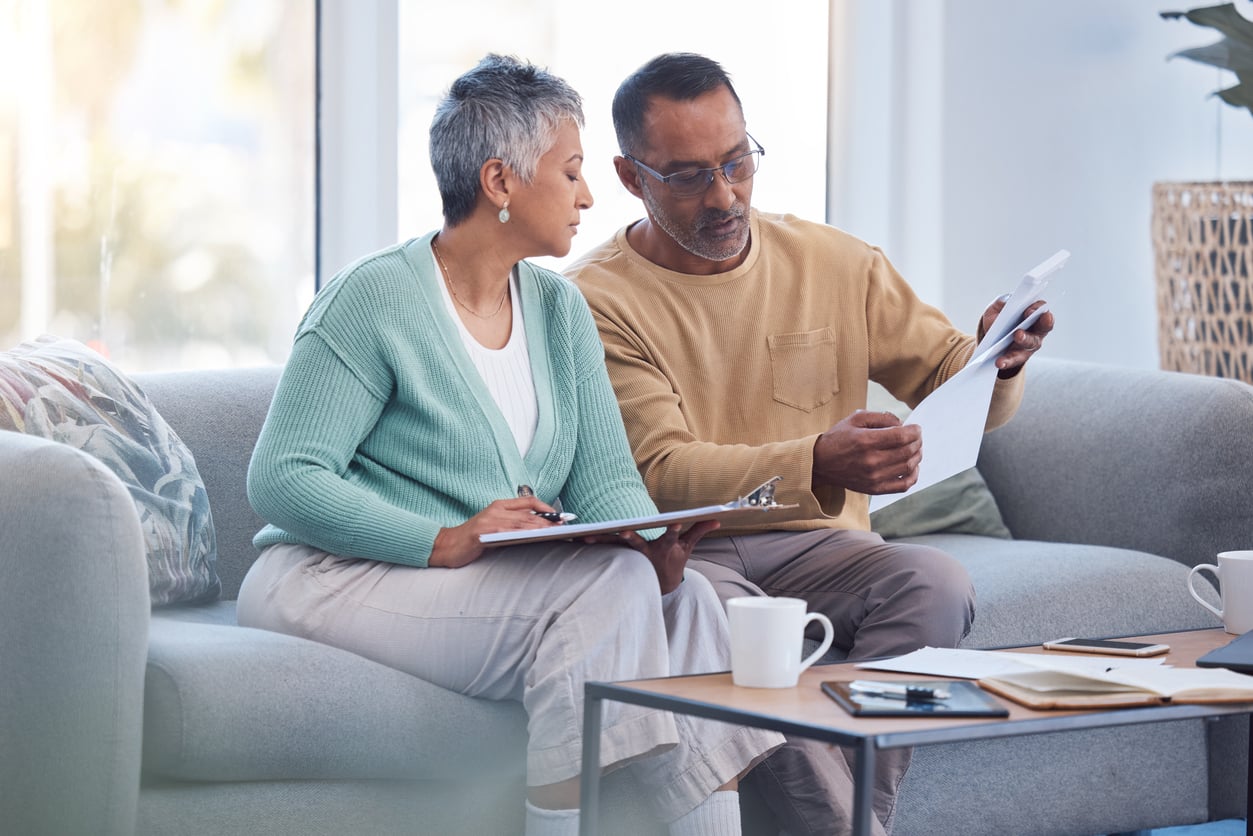 An older couple sits on a sofa, reviewing documents together. The man holds papers while the woman writes on a clipboard. There are coffee mugs, a notebook, and papers on the table in front of them.