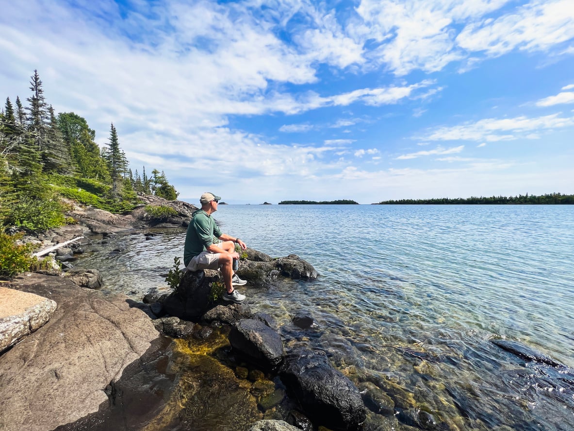 A person in a cap sits on a rocky shore, gazing over a calm, clear lake surrounded by trees under a partly cloudy blue sky.