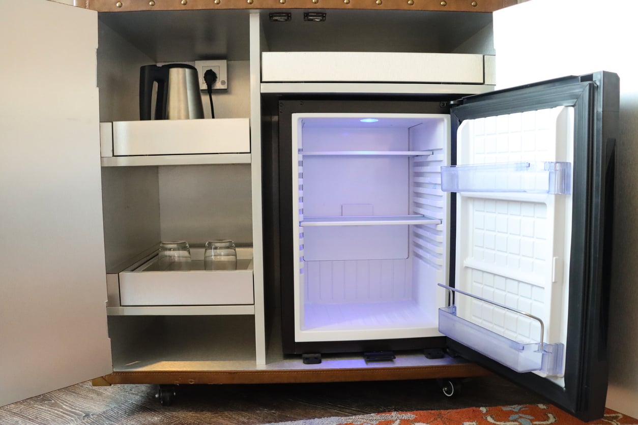 A small open fridge with empty shelves, open door, glass cups on a shelf, and an electric kettle inside a cabinet.