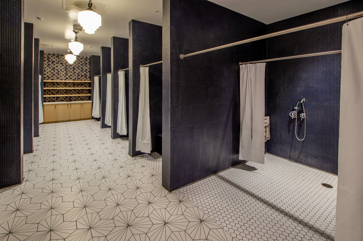 A modern shower room with dark tiled walls, several shower stalls with white curtains, white geometric floor tiles, and wooden shelves in the background.