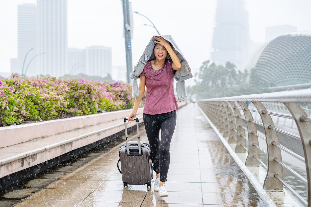 A woman walks on a rainy city sidewalk, using a jacket to shield herself—one of those classic vacation mistakes of not packing an umbrella. She pulls a rolling suitcase and is surrounded by city buildings, flowers, and wet pavement.