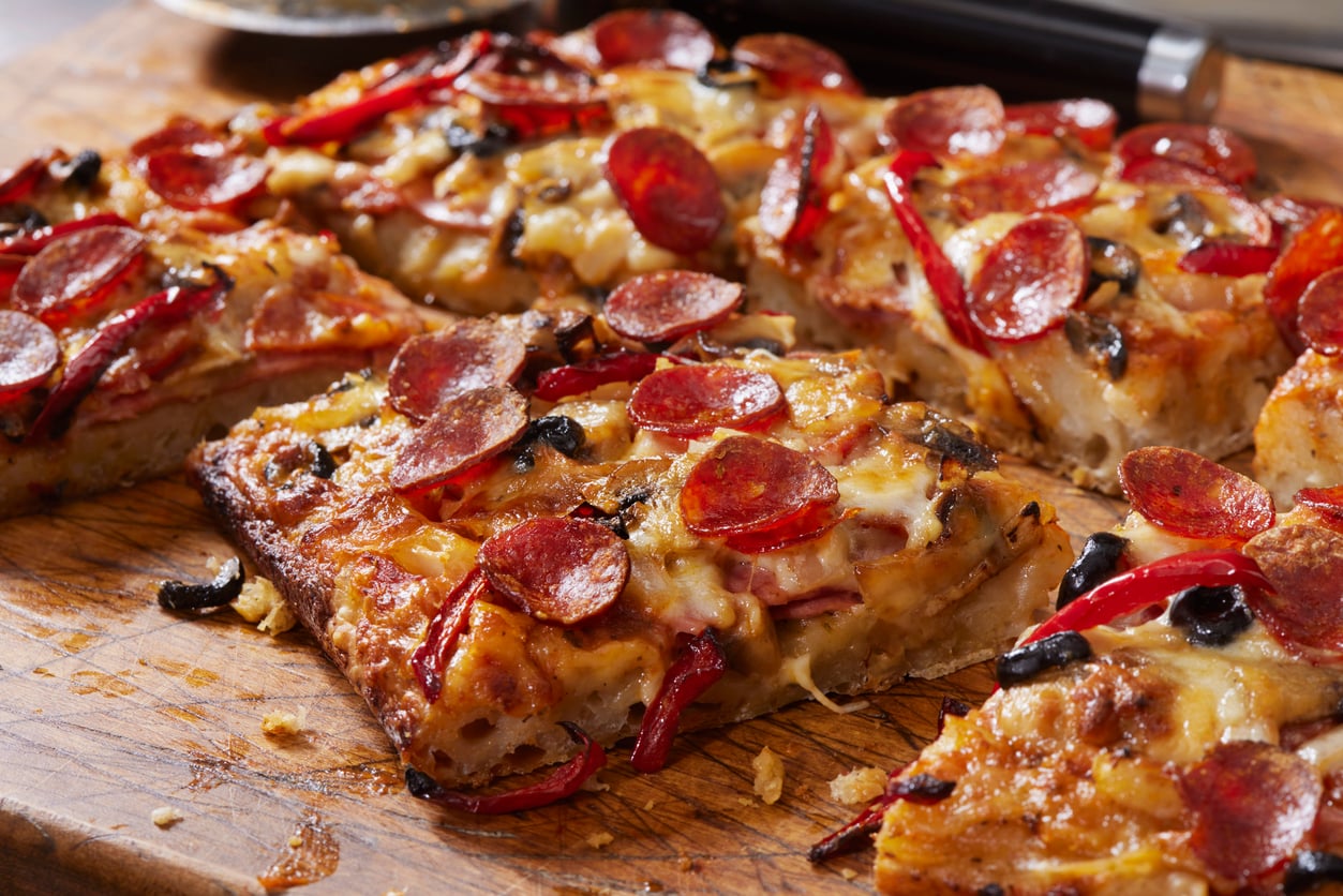 Close-up of rectangular slices of pizza topped with pepperoni, red peppers, black olives, and melted cheese on a wooden cutting board.