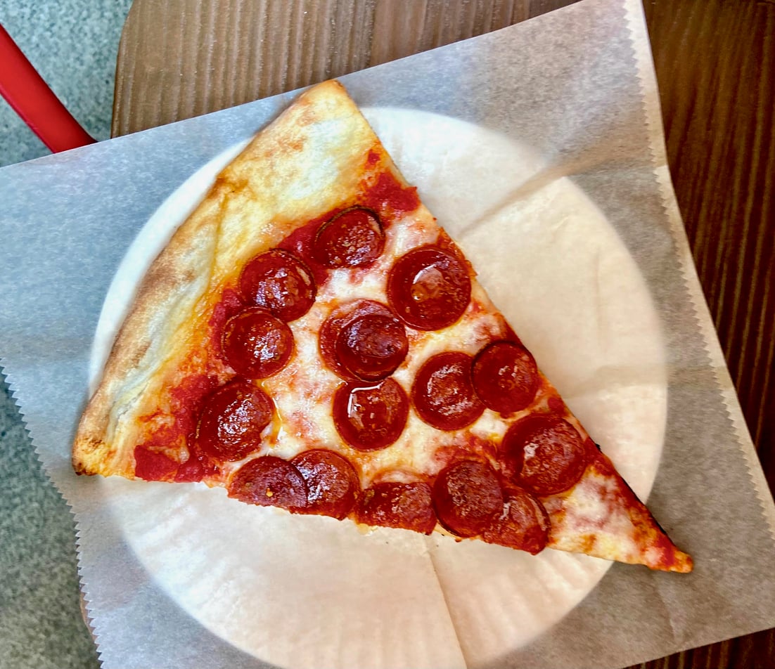 A slice of pepperoni pizza sits on a white paper plate, which is placed on a piece of parchment paper atop a wooden table.