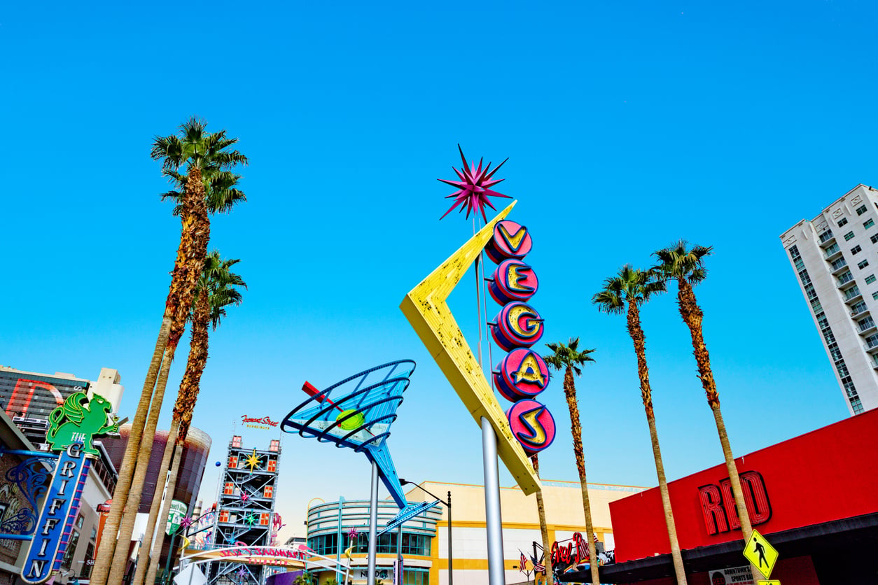 Colorful Vegas sign and a large martini glass sculpture stand amid palm trees against a clear blue sky, with nearby buildings and other neon signage visible in the background.