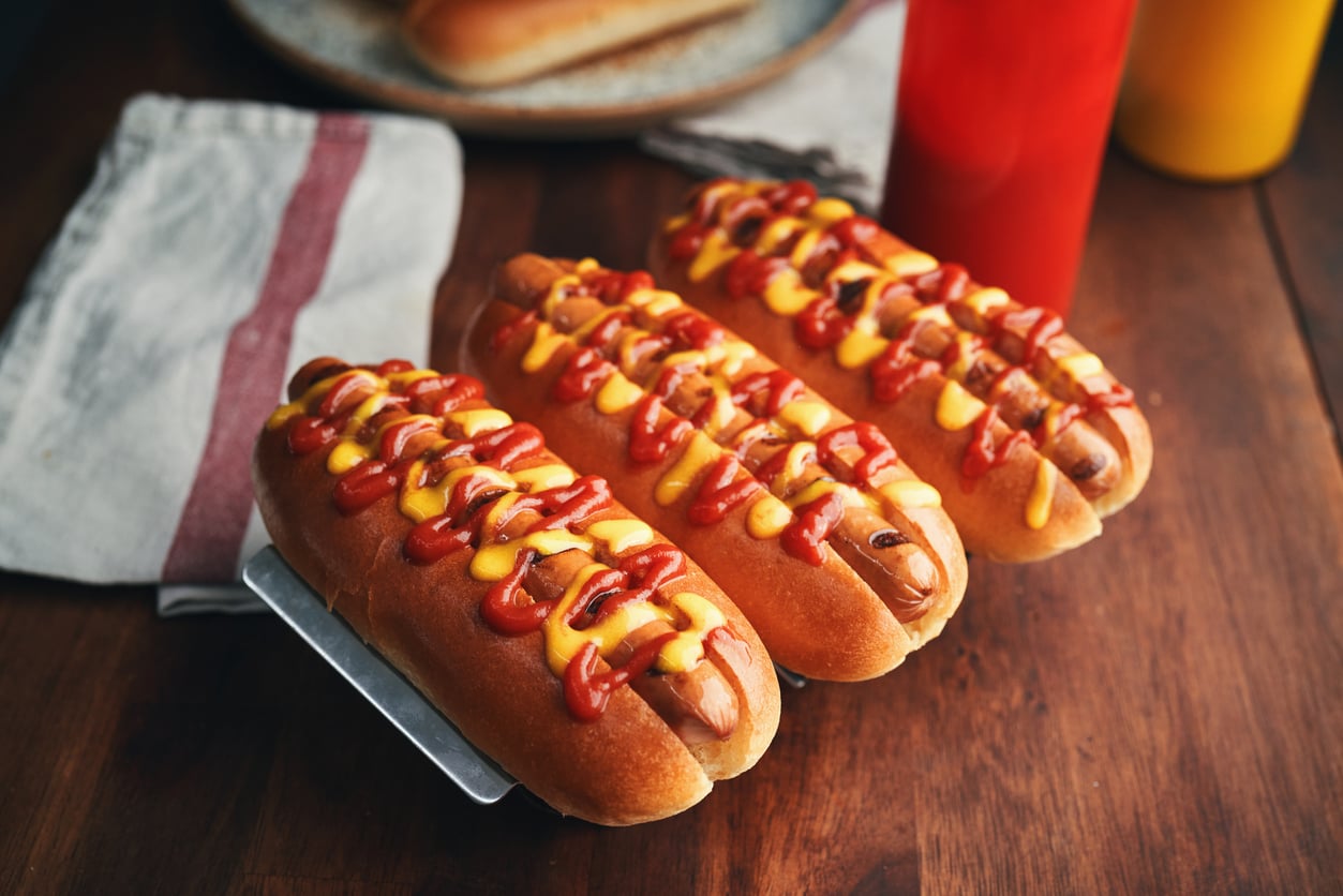 Three hot dogs in buns topped with ketchup and mustard are arranged on a wooden table next to a white cloth napkin with a red stripe. These classic foods are complemented by bottles of ketchup and mustard visible in the background.
