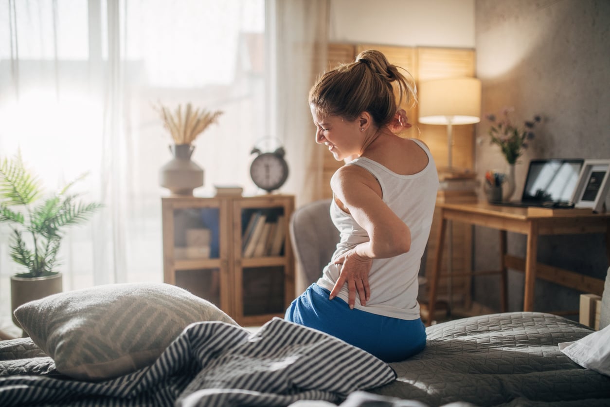 A woman sits on the edge of a bed in a sunlit bedroom, holding her lower back and appearing to be in discomfort or pain. The room is cozy, with plants, a clock, and soft lighting visible in the background.
