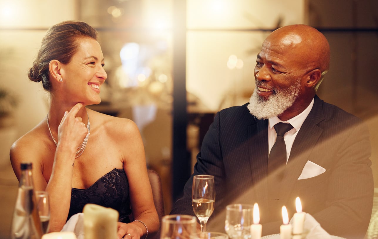 A smiling woman in a black dress and a man in a suit and tie sit together at a candlelit dinner table, enjoying each other’s company.