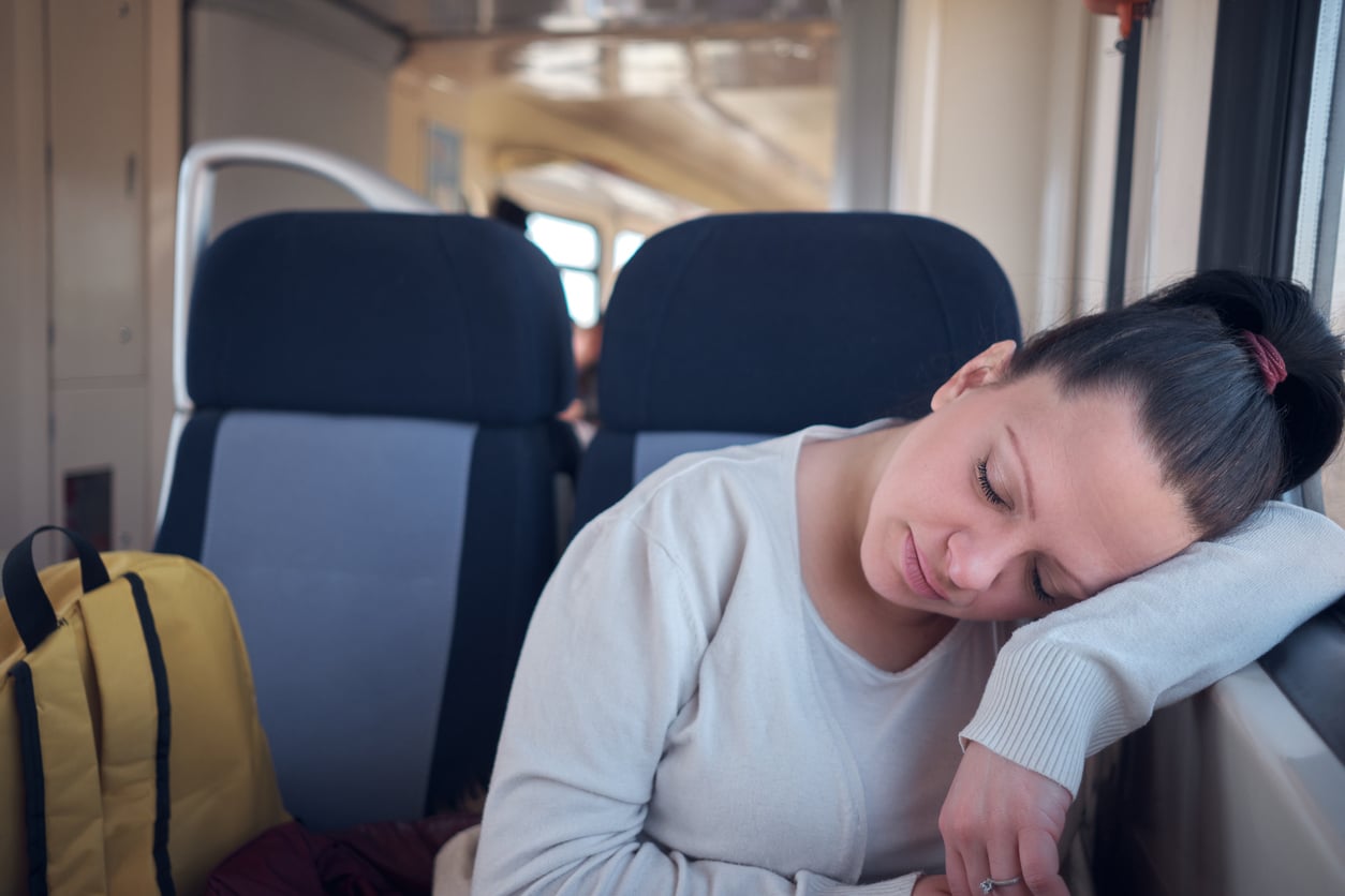 A woman with dark hair in a ponytail rests her head on her arm against a train window, eyes closed, wearing a light sweater. A yellow backpack sits on the seat beside her.