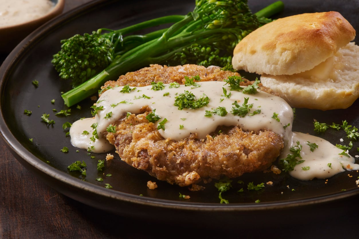 A plate with chicken fried steak topped with white gravy and parsley, served with broccolini and a biscuit.