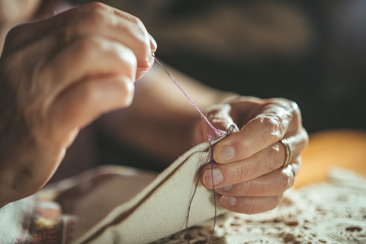 Close-up of hands sewing fabric with a needle and purple thread, showing careful stitching and attention to detail in a well-lit, cozy setting.