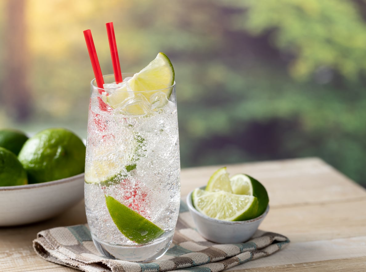 A glass of sparkling water with ice, lime wedges, and two red straws sits on a cloth napkin—an elegant touch reminiscent of everyday foods that used to be considered fancy. Nearby, a small bowl and a larger bowl filled with limes add to the fresh, vibrant scene set against a blurred green background outdoors.