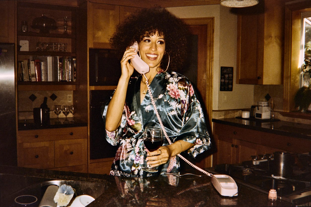 A person in a floral robe stands in a kitchen, smiling while talking on a pink corded phone and holding a glass of red wine.