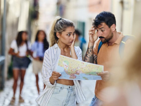 A woman and man stand in a cobblestone street, looking confused while reading a map&mdash;caught in the midst of classic vacation mistakes. The woman is touching her head and the man is scratching his forehead as they try to get their bearings, while blurry people walk by in the background.