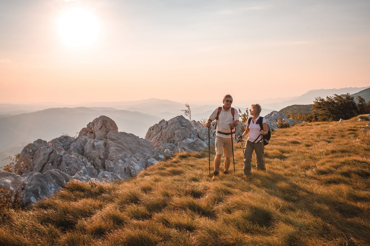 Two older adults enjoying unforgettable vacation experiences as they hike on a grassy, sunlit mountain ridge at sunset, surrounded by rocks, with distant hills visible under a soft orange sky.
