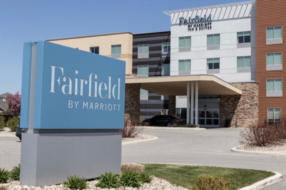 A Fairfield by Marriott hotel exterior, part of one of the renowned hotel chains, with a prominent blue sign in front, a modern building, a covered entrance, and landscaped surroundings on a sunny day.
