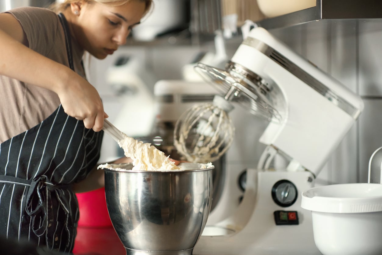 A person in an apron mixes batter in a metal bowl using a spatula, next to a stand mixer in a kitchen.