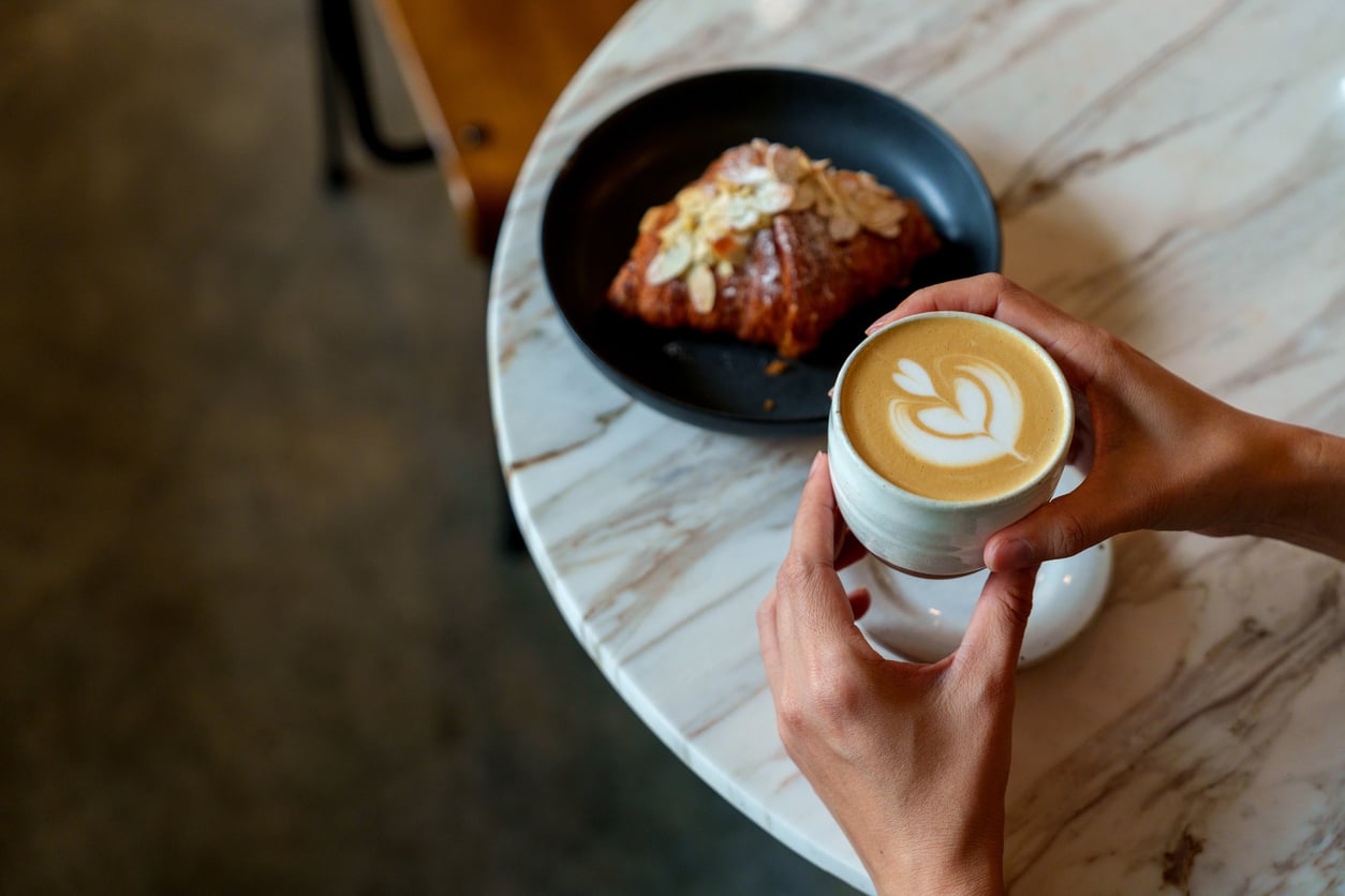 A person holds a cup of latte with heart-shaped foam art at a marble table, next to a black plate with an almond croissant.