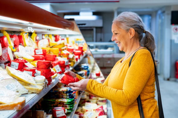 A woman in a yellow sweater smiles while selecting a red-wrapped cheese from a display case filled with various cheeses at a grocery store.