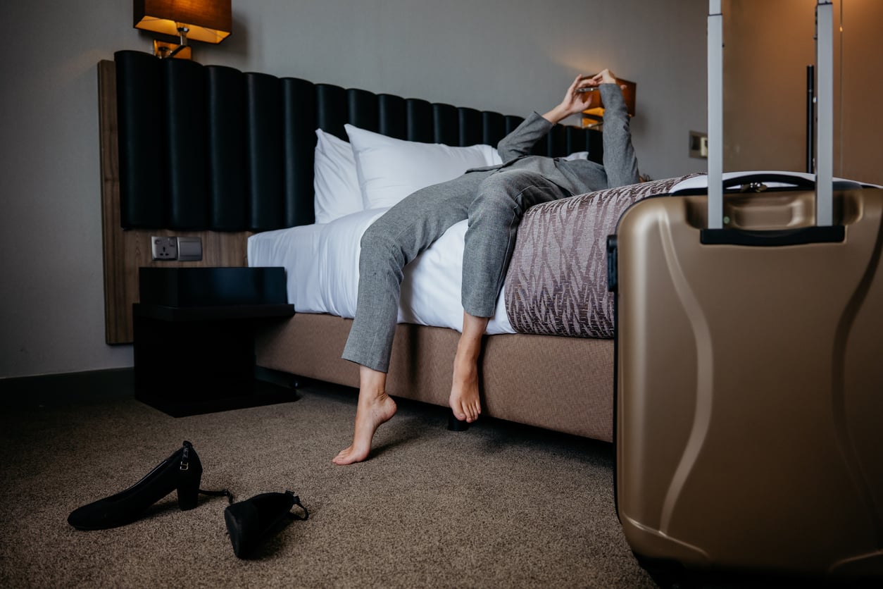 A person in gray clothes lies sprawled on a hotel bed, feet on the floor, with shoes beside the bed and a suitcase in the foreground. The person appears to be using a phone.
