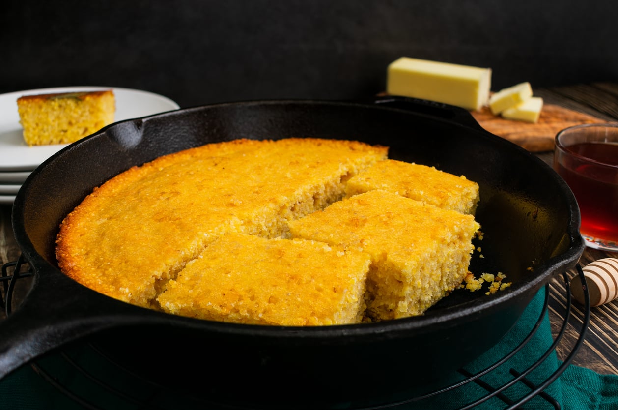 A skillet of golden cornbread with square pieces cut, one slice missing. In the background, there’s butter on a wooden board, a glass of dark liquid, and a plate with a cornbread slice.