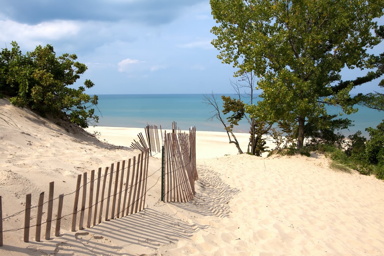 Sandy path with a wooden fence leads to a beach, bordered by trees and green bushes, with calm blue water and a partly cloudy sky in the background.