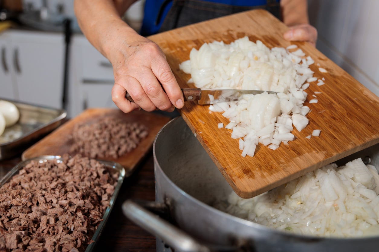 A person transfers chopped onions from a wooden cutting board into a large pot, with cooked, diced meat visible nearby on trays in a kitchen setting.