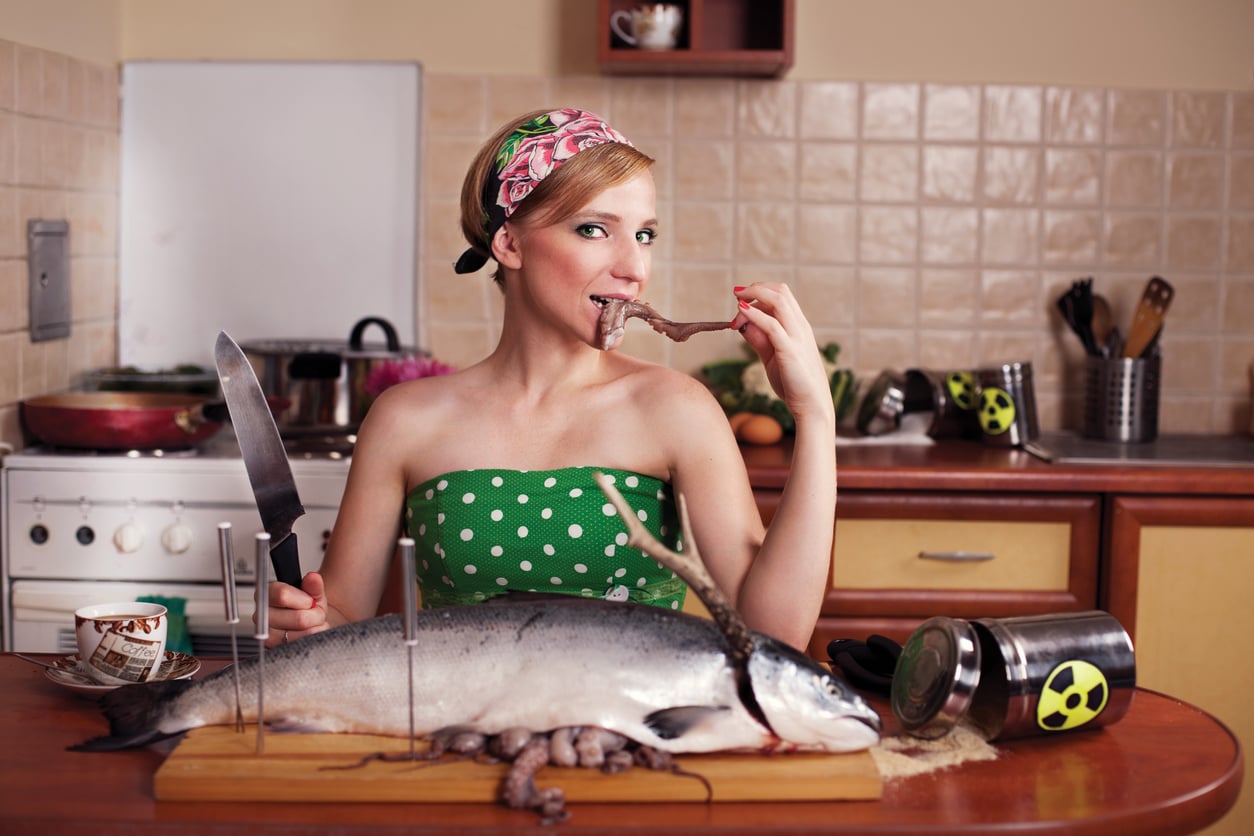 A woman in a green polka dot dress sits in a kitchen, holding a knife and eating an octopus tentacle. A large fish and kitchen tools with radioactive symbols are on the counter in front of her.